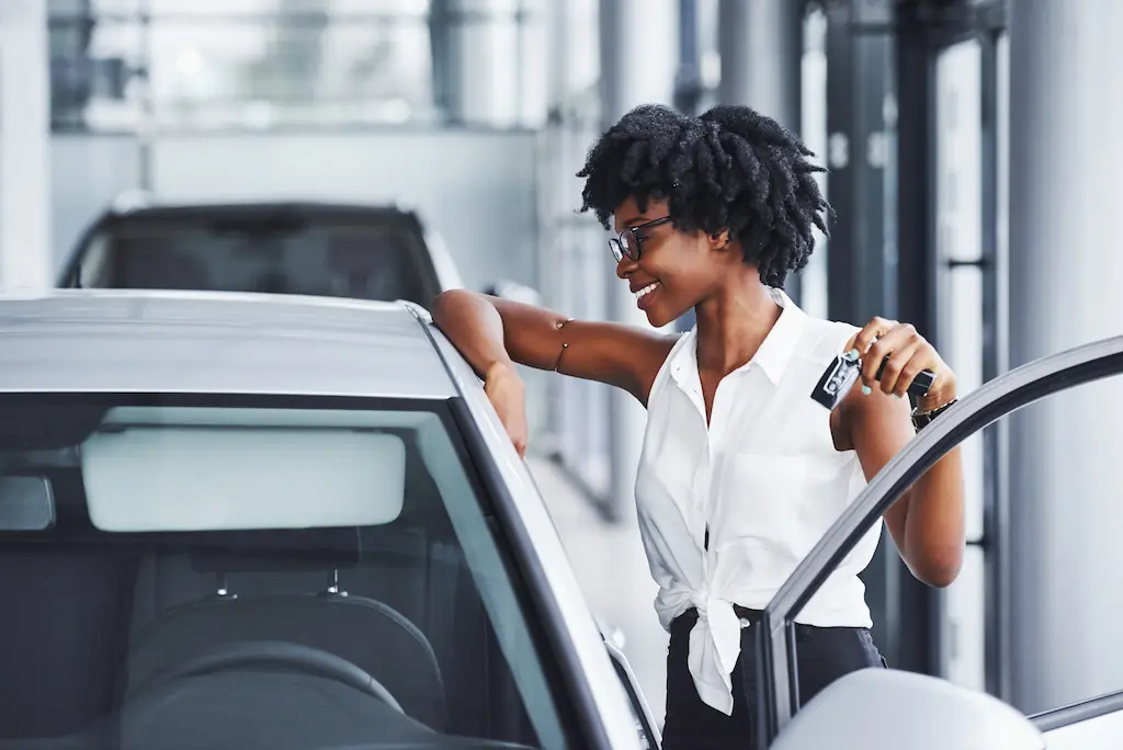 Mulher sorrindo apoiada na lateral de um carro com a chave em mãos.