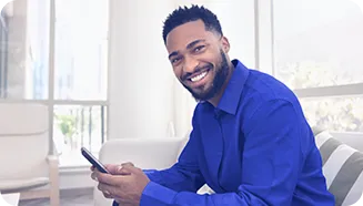 Homem sorridente, com camisa azul, segurando um celular. Ele está sentado em um ambiente iluminado.