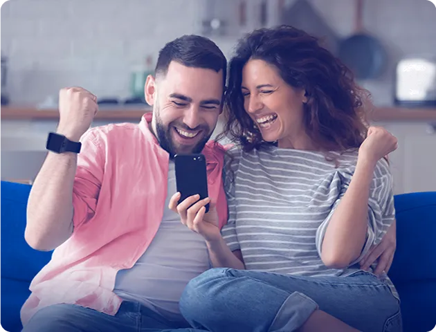 Na imagem, temos um homem e uma mulher sentados no sofá felizes, ambos olhando para o celular que a mulher está segurando.