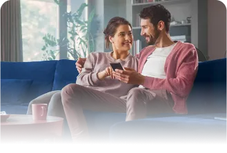 Um casal formado por um homem e uma mulher sentados em um sofá se olhando e mexendo em um celular.