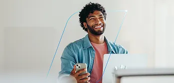 Homem sorrindo, segura um celular e usa um notebook em um ambiente interno com fundo claro.