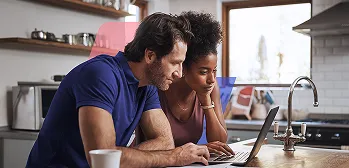 Um homem e uma mulher olham juntos para a tela de um notebook em uma cozinha.