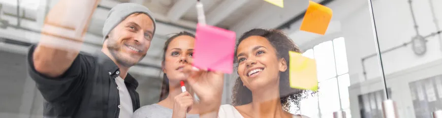 Duas mulheres e um homem em frente a um quadro transparente realizando anotações em post-its.