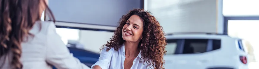 Duas mulheres em uma concessionária. Uma mulher de cabelo cacheado sorri ao apertar a mão da outra.