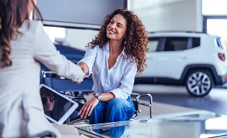 Duas mulheres em uma concessionária. Uma mulher de cabelo cacheado sorri ao apertar a mão da outra.