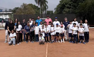 Várias pessoas juntas, adultos, crianças e jovens em uma quadra de tênis, segurando raquetes, posicionados para uma foto.
