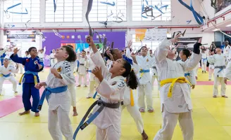 Várias crianças com quimonos e faixas de jiu-jitsu, em um tatame, comemorando felizes.