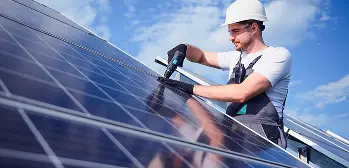 Técnico com capacete instala painéis solares sob céu azul, ele está usando ferramenta e equipamentos de segurança.