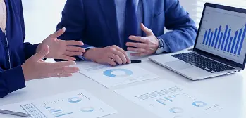 Duas pessoas em reunião de negócios revisando gráficos e relatórios financeiros sobre a mesa, com laptop aberto.