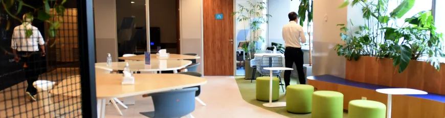 Ambiente de escritório moderno com mesas, assentos verdes, plantas e pessoas ao fundo.