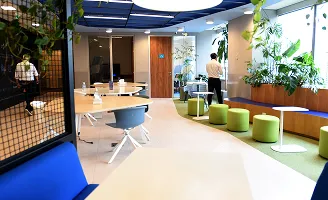 Ambiente de escritório moderno com mesas, assentos verdes, plantas e pessoas ao fundo.