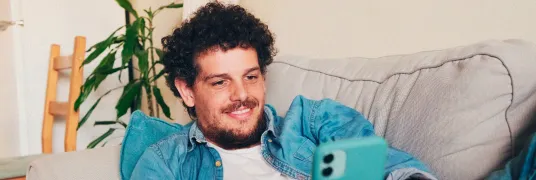 Homem sorridente, deitado em um sofá enquanto mexe em um celular.