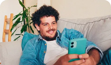Homem sorridente, deitado em um sofá enquanto mexe em um celular.
