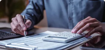 Pessoa com documentos, computador e calculadora em uma mesa.