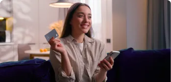 Mulher sorrindo com o cartão BV em uma das mãos e na outra um celular.