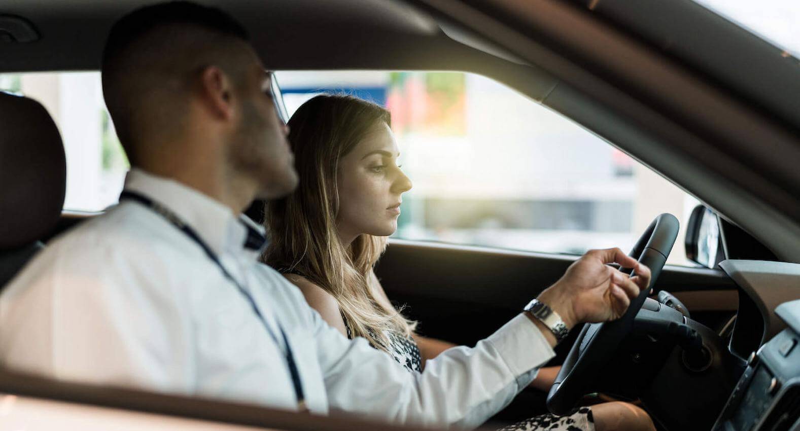 A imagem mostra um atendente de uma concessionária, dentro de um carro, com uma mulher na direção.