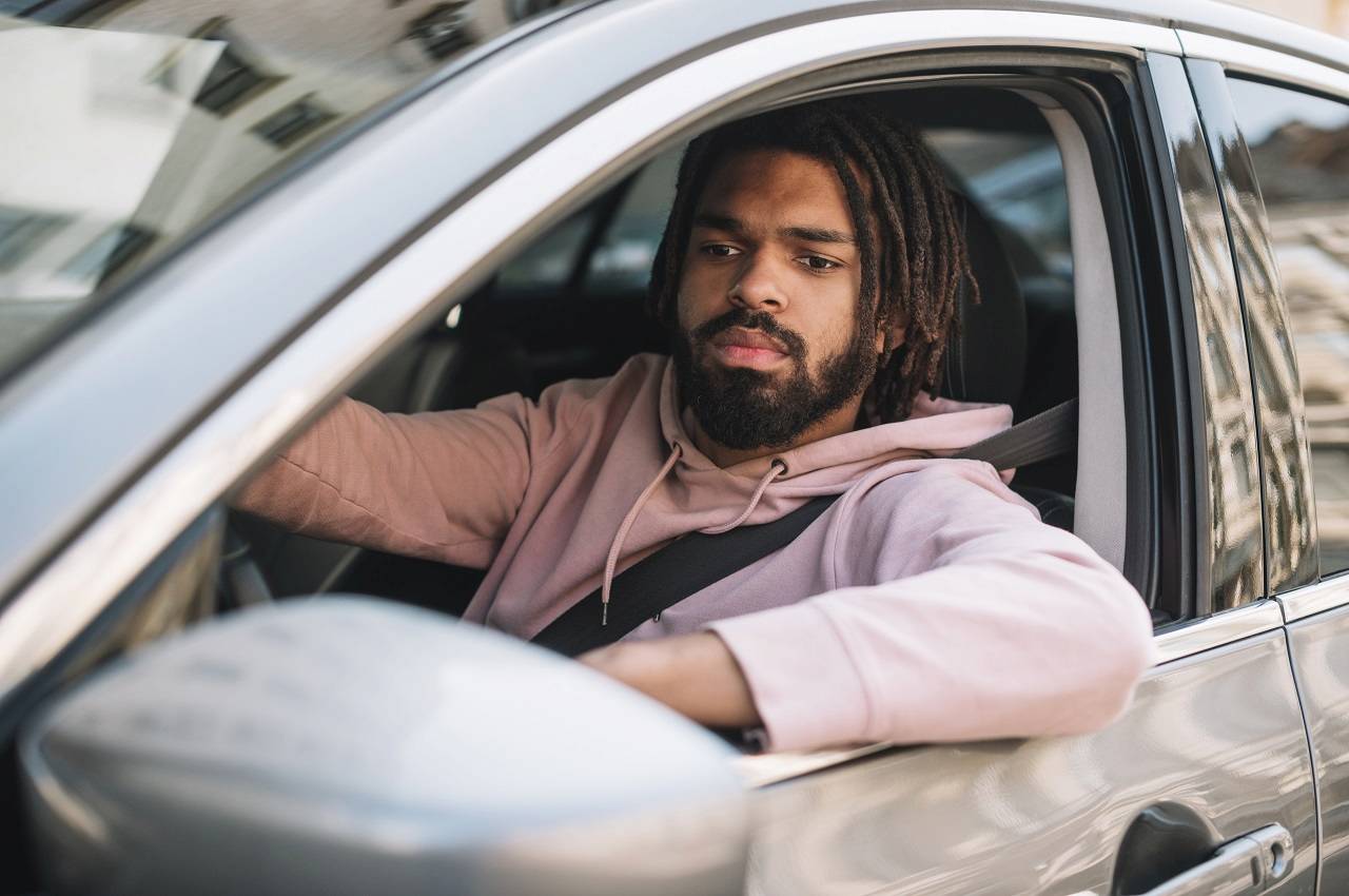 Homem de barba e tranças, com expressão concentrada e dirigindo um veículo prata.