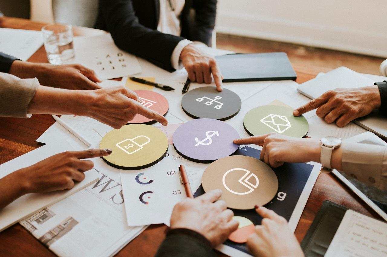 A imagem mostra uma mesa em uma reunião de alinhamento. A imagem mostra apenas as mãos das pessoas e alguns papéis com blocos de ícones financeiros em cima.