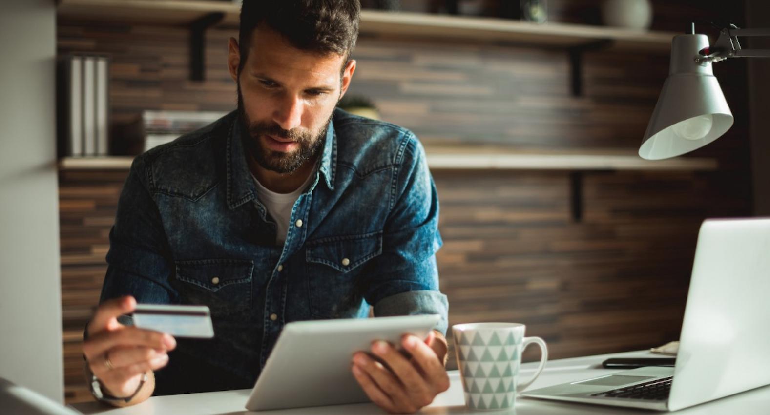 A imagem mostra um homem usando seu tablet e seu cartão de crédito.