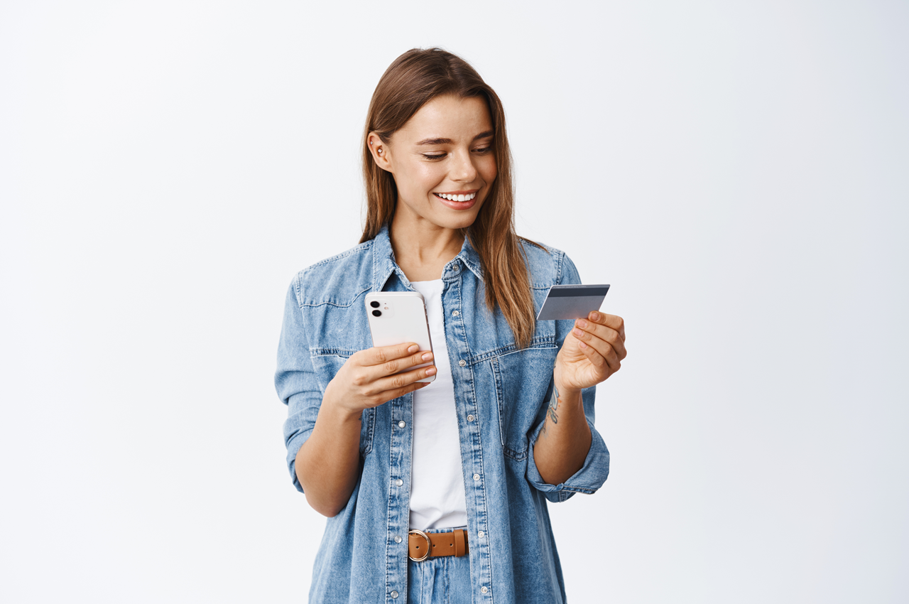 Mulher sorrindo enquanto segura um smartphone em uma mão e um cartão de crédito na outra. Ela está vestindo uma jaqueta jeans sobre uma camiseta branca, e seus cabelos são longos e ondulados. O fundo é liso e de cor clara.