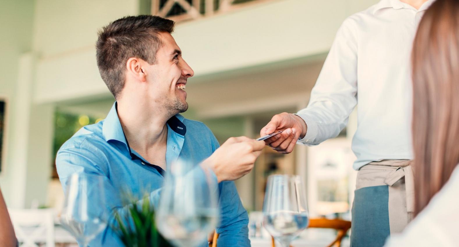 A imagem mostra um homem em um restaurante, realizando o pagamento com seu cartão de crédito.