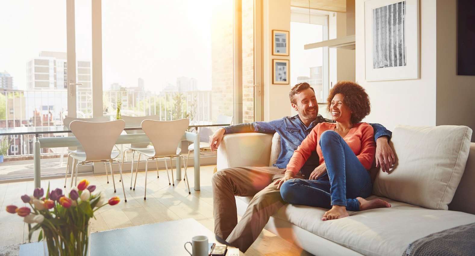 A imagem mostra um casal sorridente, sentados no sofá de sua casa.