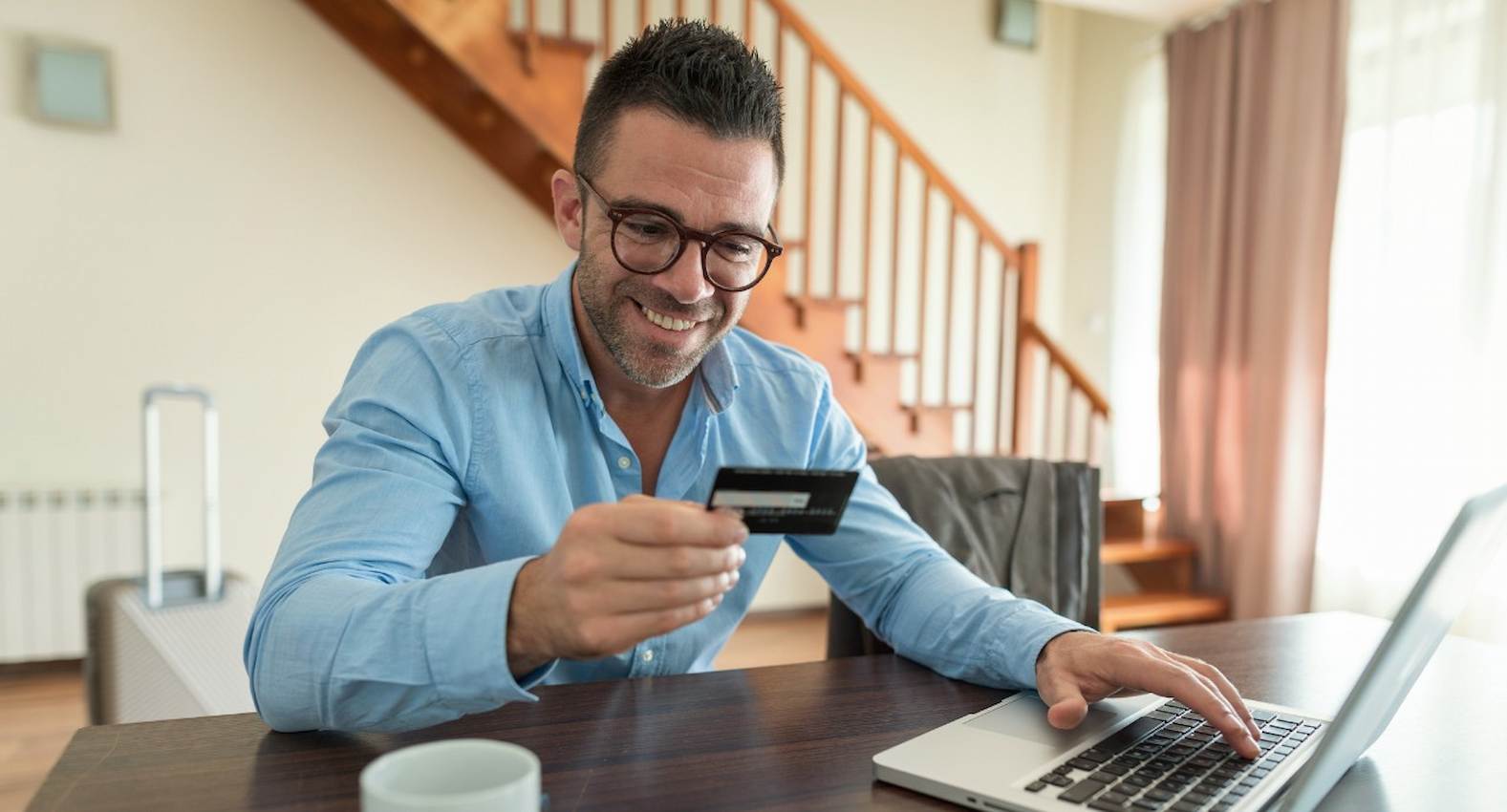 A imagem mostra um homem sorridente, usando seu cartão de crédito para fazer compras em seu laptop.