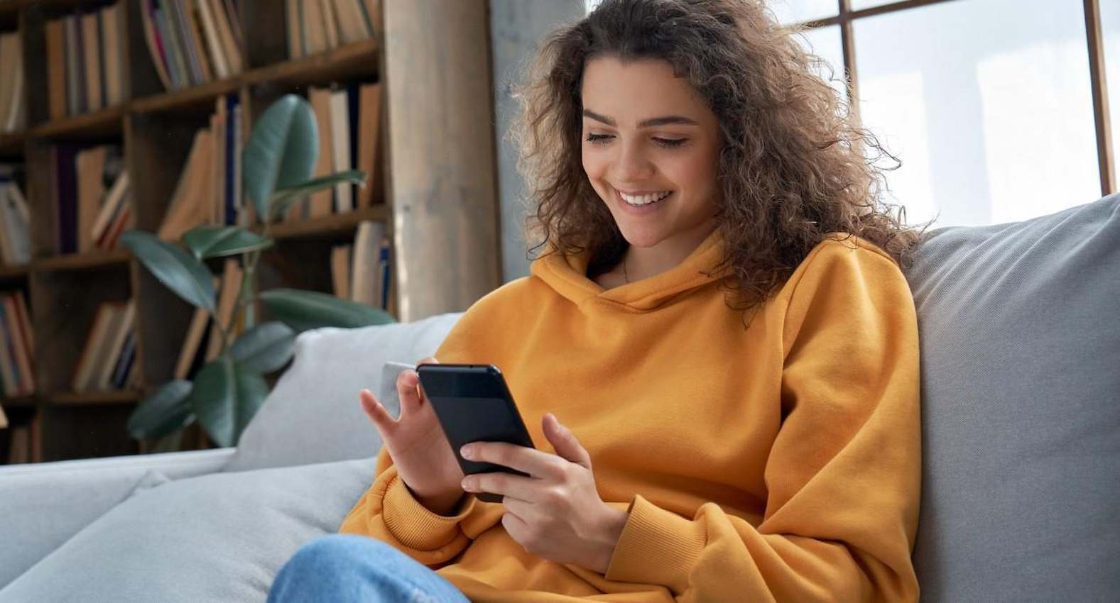 A imagem mostra uma mulher, vestindo uma blusa de frio laranja, usando seu celular. Ela está sentada em um sofá e sorridente.