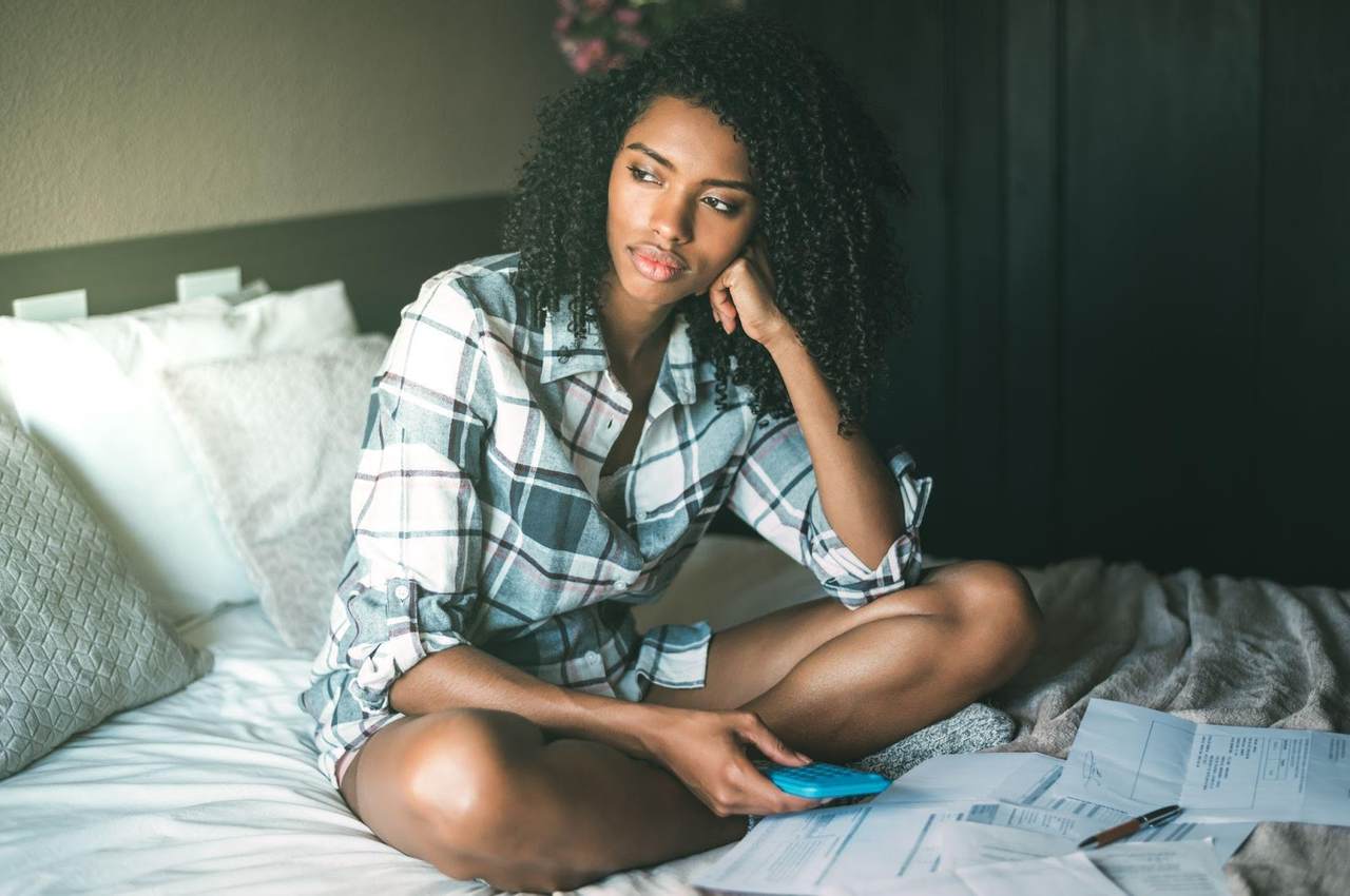 A imagem mostra uma mulher, sentada à sua cama, com alguns boletos em volta. Ela está com uma calculadora na mão e aparenta estar preocupada.