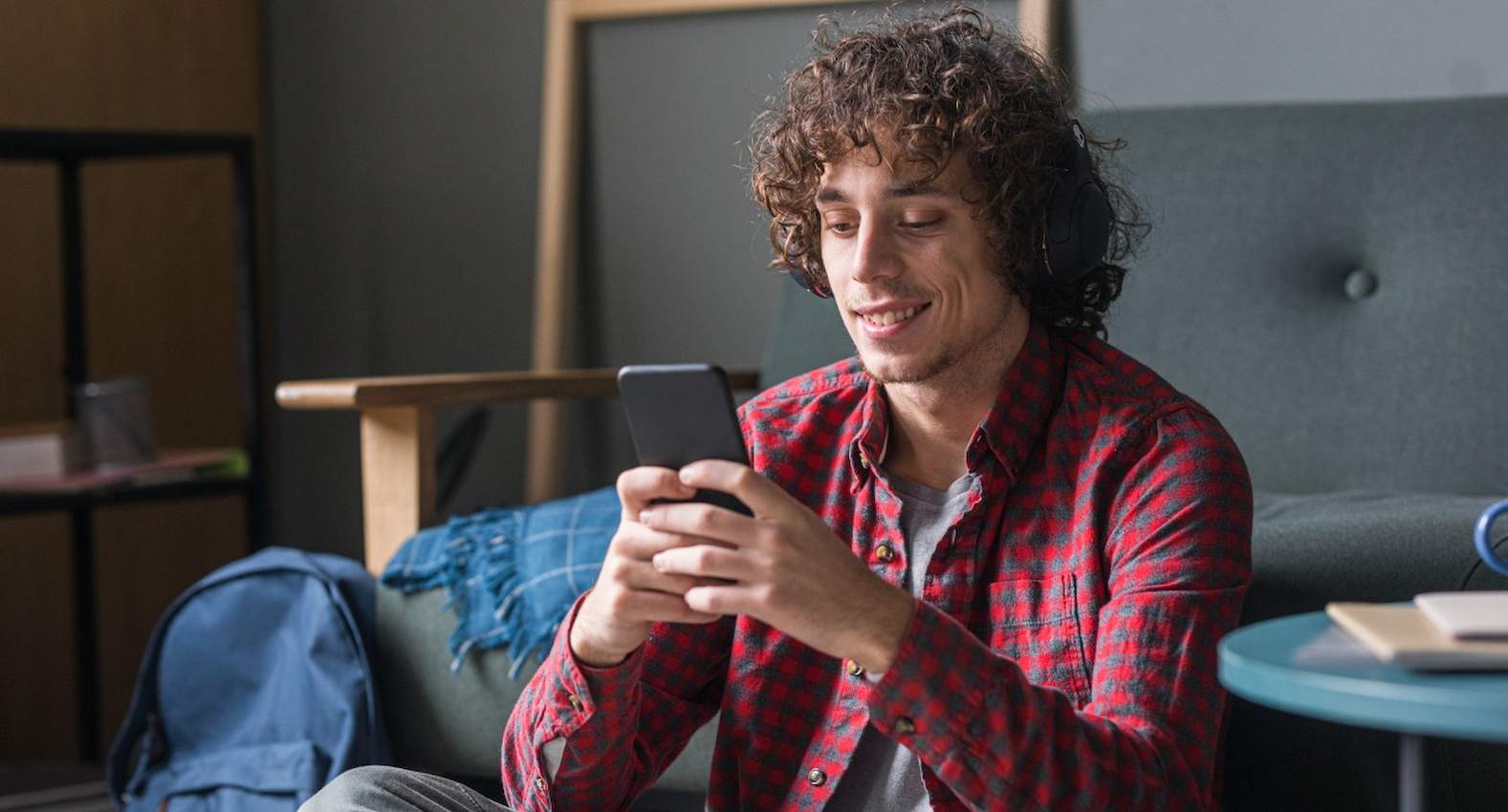 A imagem mostra um homem, usando camisa xadrez e fones de ouvido, usando seu celular. Ele está sorridente.