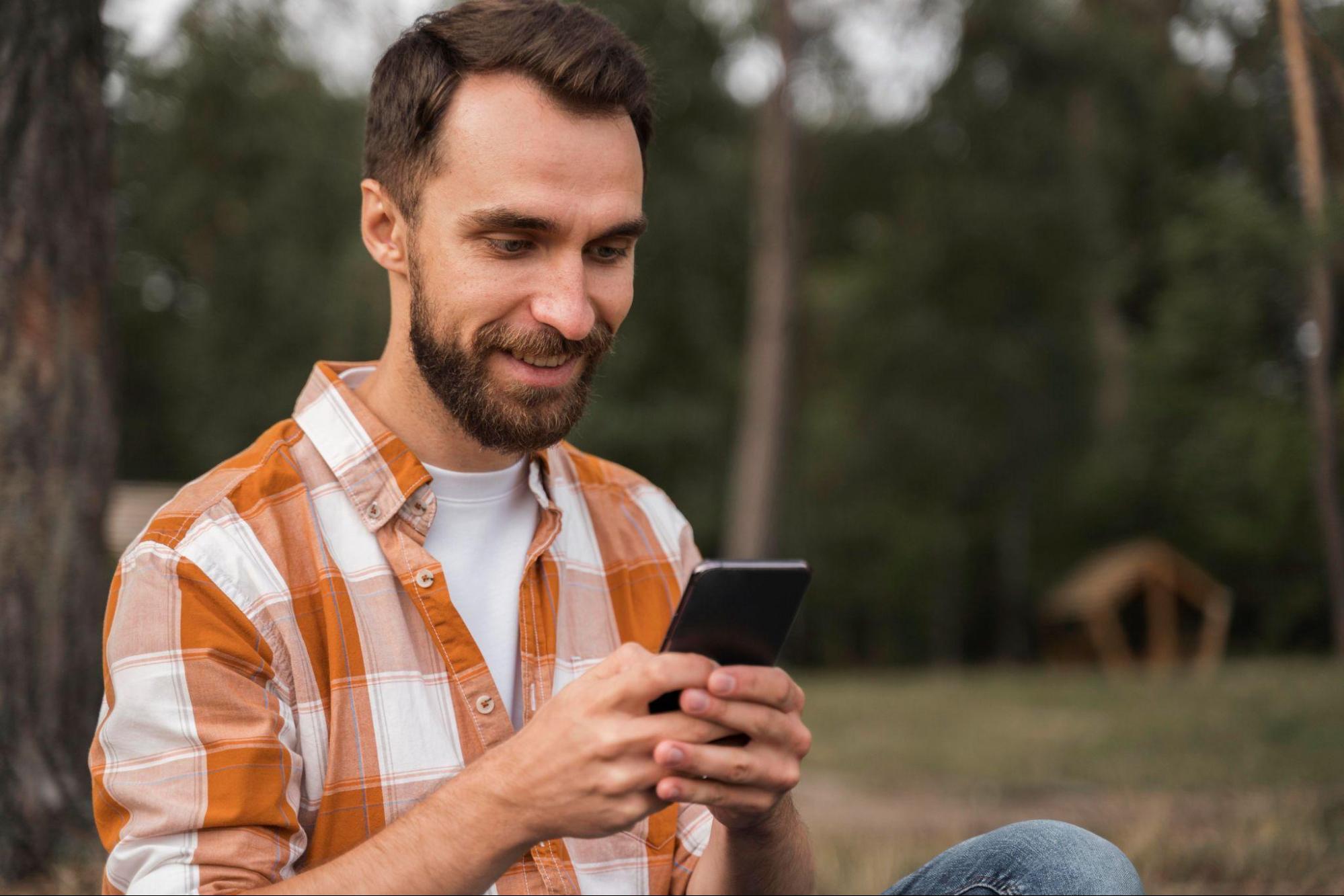 Homem sorridente com barba, usando uma camisa xadrez laranja, segurando um smartphone. Ele está ao ar livre, próximo a árvores.