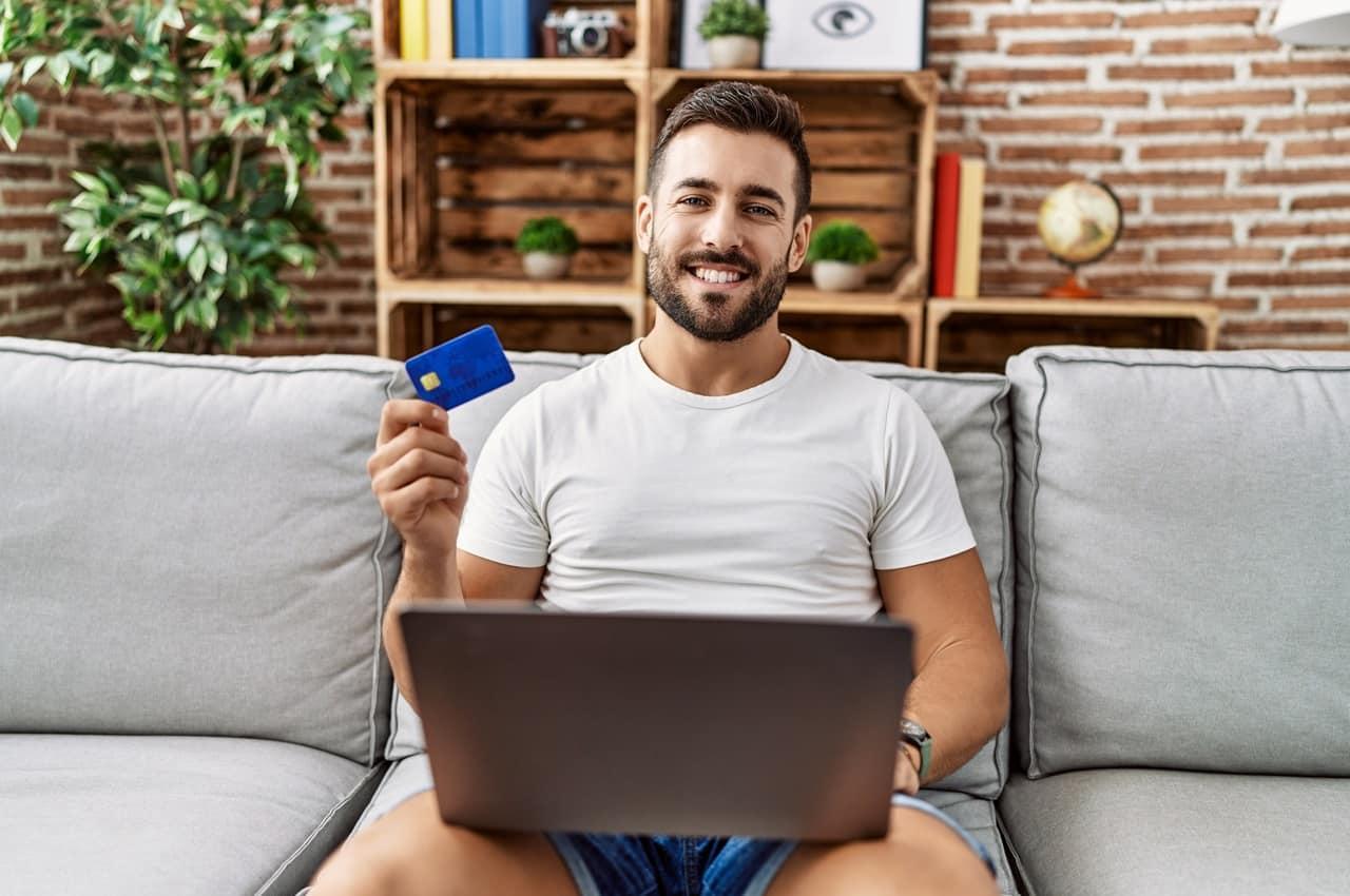 Homem sorrindo enquanto segura um cartão de crédito em uma mão e um laptop aberto na outra. Ele está sentado em um sofá cinza e usa uma camiseta branca. Ao fundo, há prateleiras de madeira com plantas e objetos decorativos. 