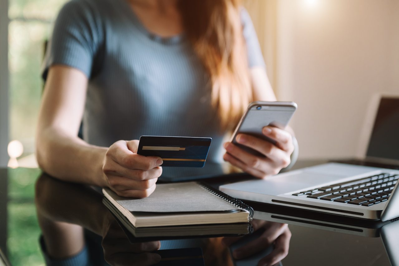 A imagem mostra uma mulher fazendo compras com o cartão de crédito em seu celular. Em sua mesa, há um laptop e um caderno.