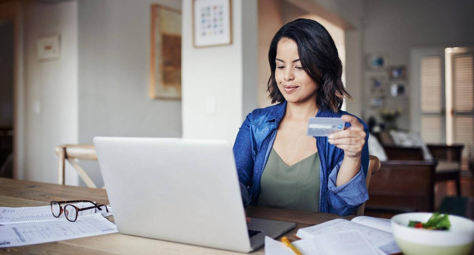 A imagem mostra uma mulher usando seu laptop e fazendo compras no cartão de crédito. Em cima da mesa, há alguns papéis e um óculos de grau.