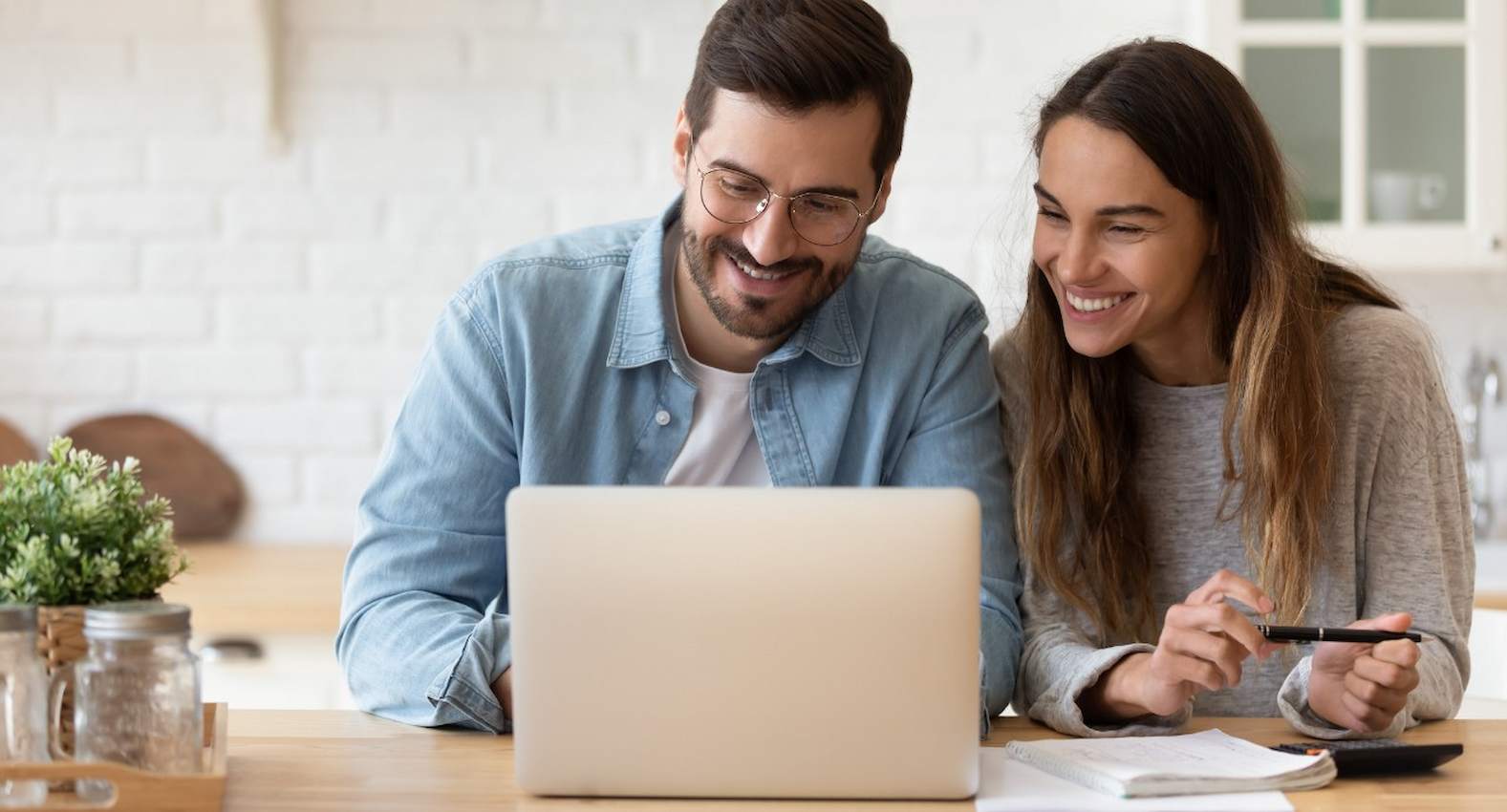 A imagem mostra um casal sorridente usando um laptop. A mulher está com uma caneta na mão.