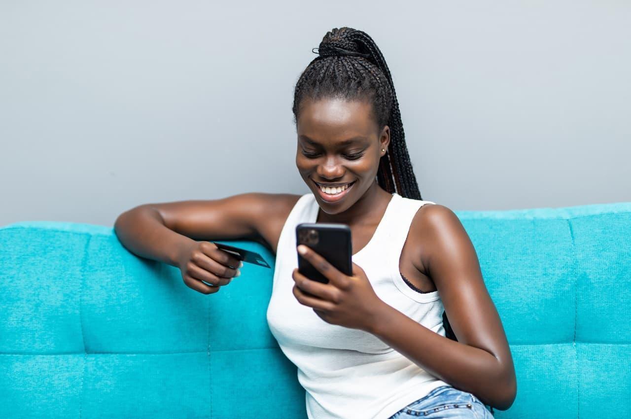 Mulher sorridente vestindo blusa básica branca e calça jeans, sentada em sofá azul e segurando um cartão de crédito para pagar à vista ou parcelado no celular.