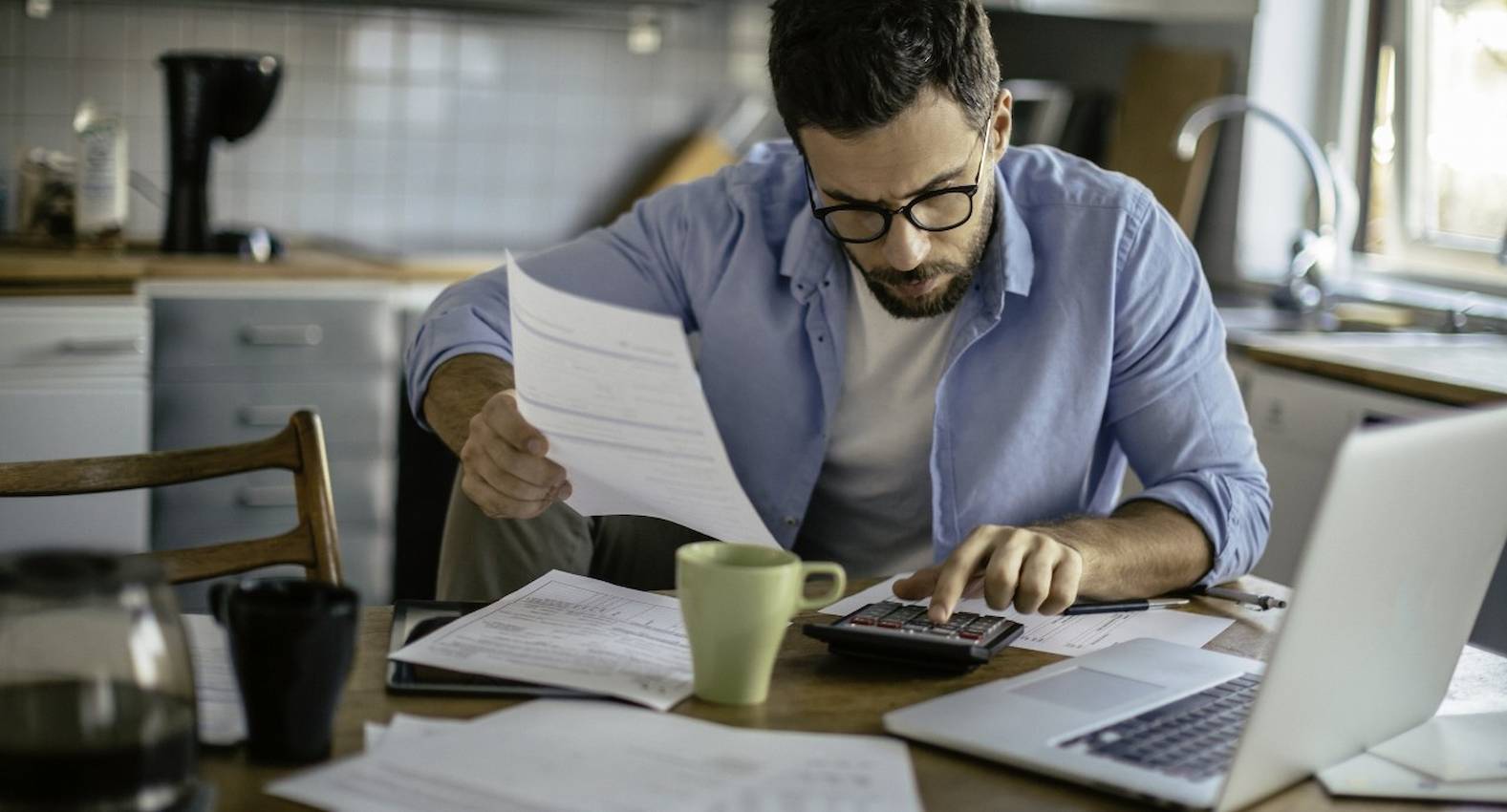 A imagem mostra um homem sentado à uma mesa com alguns papéis e uma cafeteira. Ele está realizando contas na calculadora e com o laptop ligado.