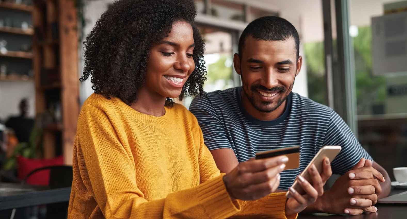 A imagem mostra um casal sorridente, sentado em um estabelecimento. A mulher está com o celular e o cartão de crédito nas mãos.