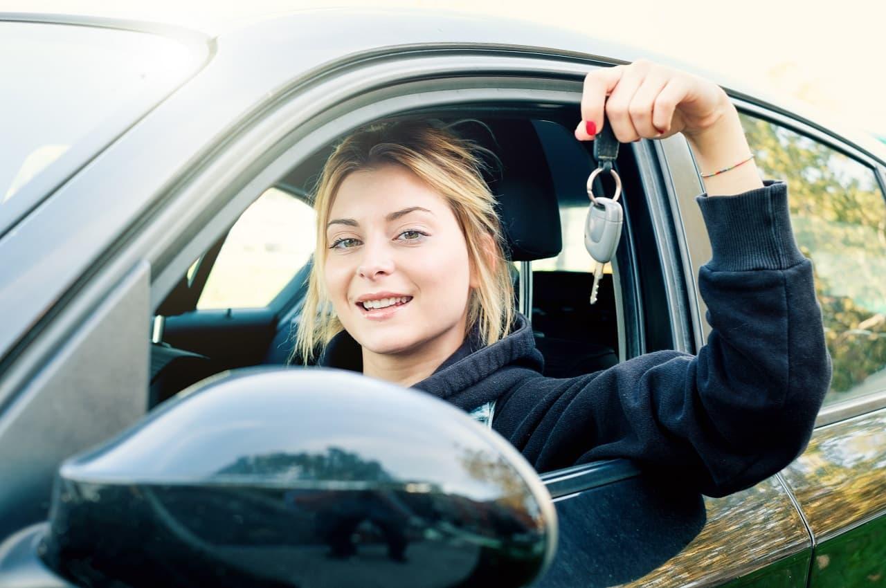 Mulher loira de olhos verdes com expressão sorridente. Ela segura uma chave de carro na mão e dirige um automóvel preto.