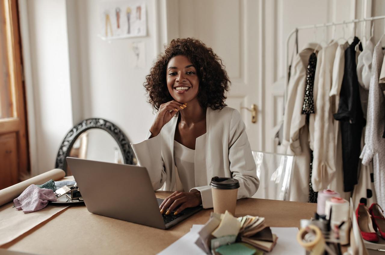 Mulher negra com expressão sorridente, sentada em uma mesa em seu escritório de moda. Ela veste blazer branco e digita no computador.