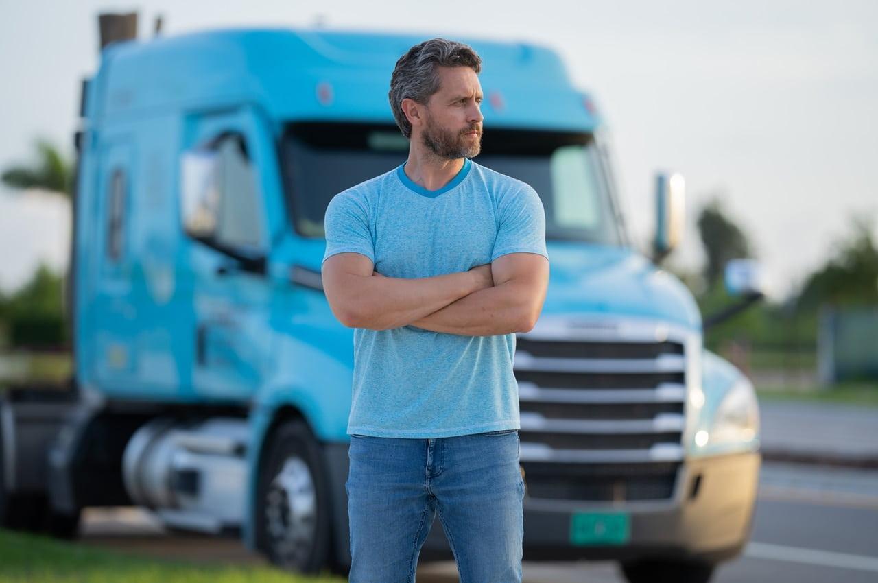 Homem de camiseta e calça jeans olhando para o lado. Ele está de braços cruzados e, ao fundo, há um caminhão azul.