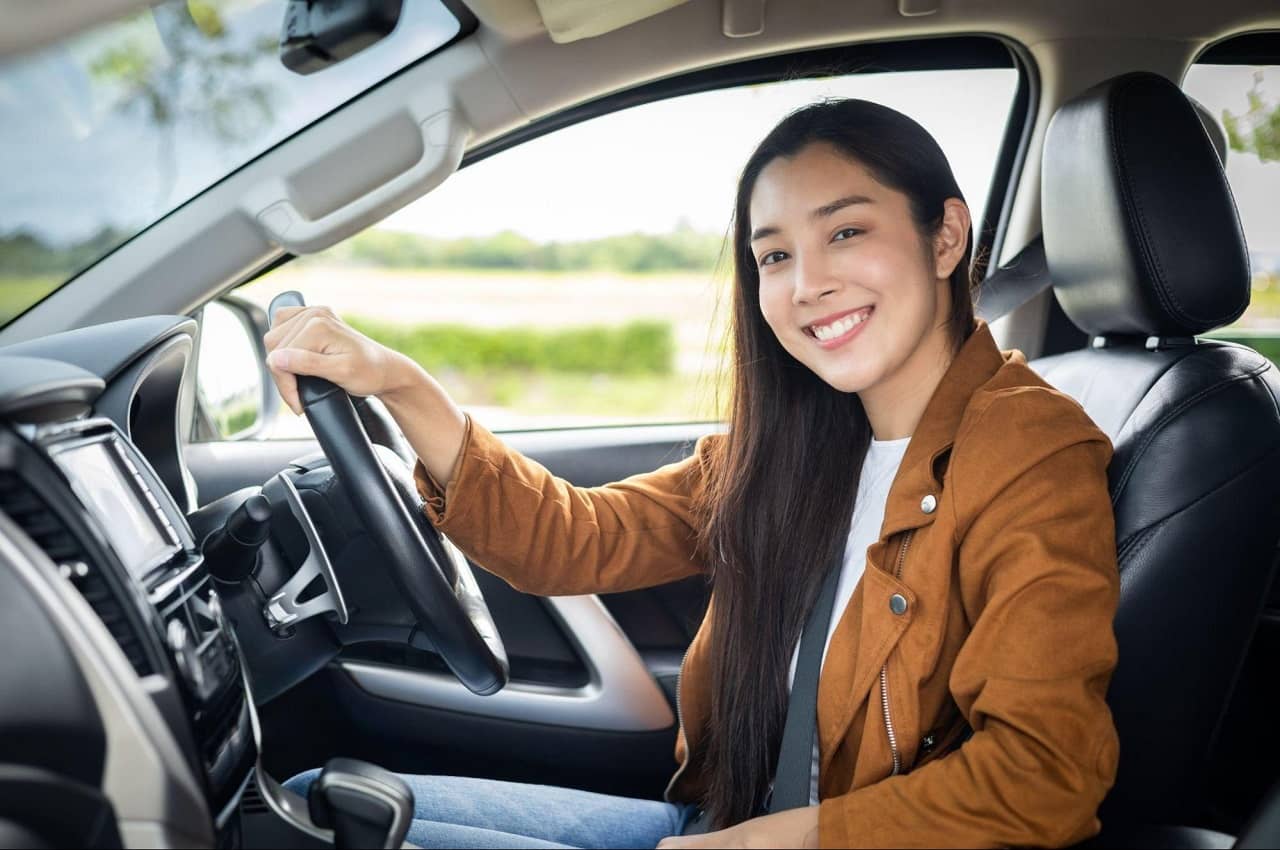 Mulher de cabelos castanhos longos, expressão sorridente e vestindo uma jaqueta de sarja marrom. Ela está sentada no banco de motorista e seguro o volante de um carro.