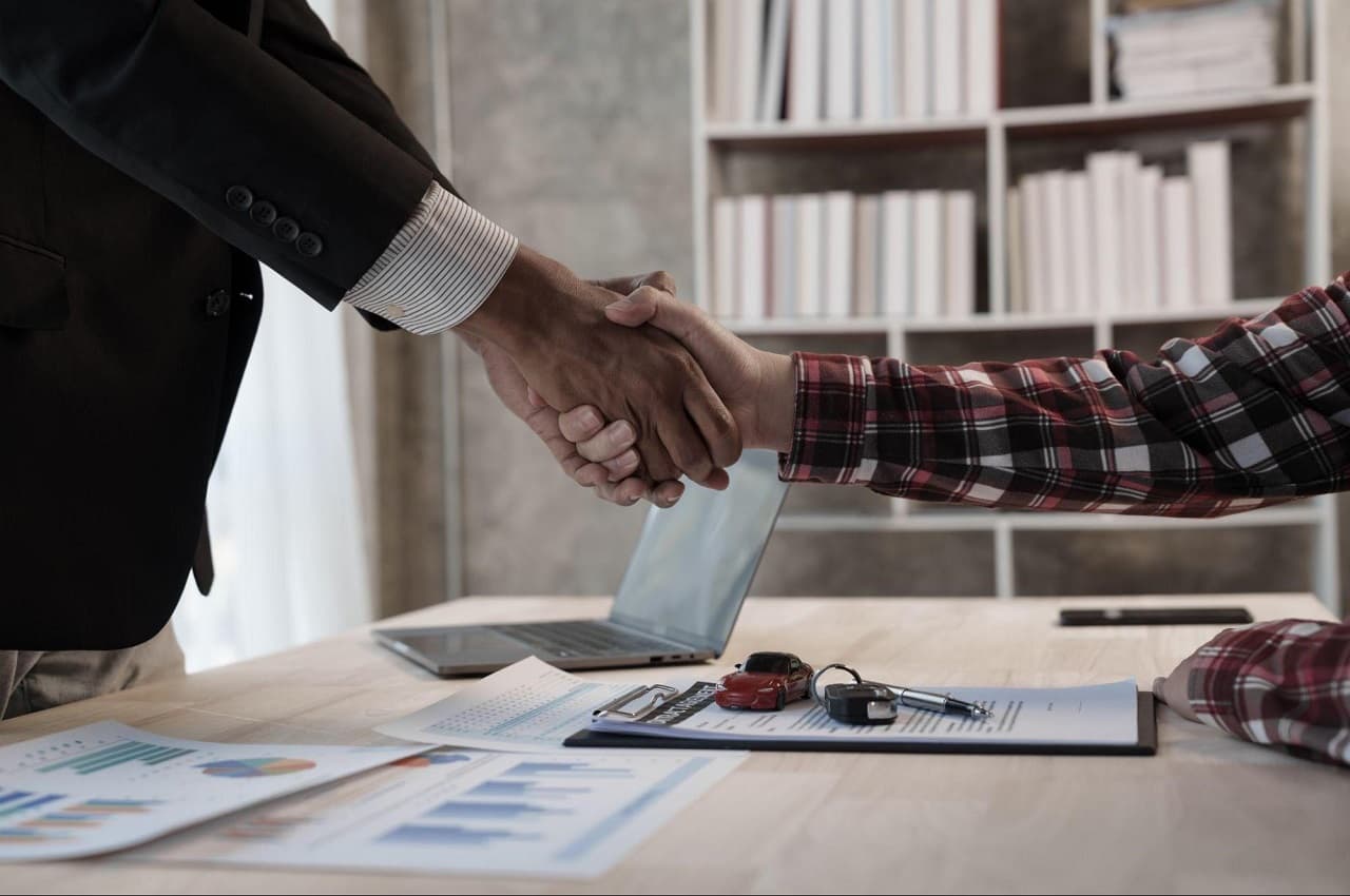 Aperto de mão entre duas pessoas após o empréstimo com garantia de veículo. Na mesa tem um contrato e em cima dele uma chave de carro e uma miniatura de um veículo vermelho.