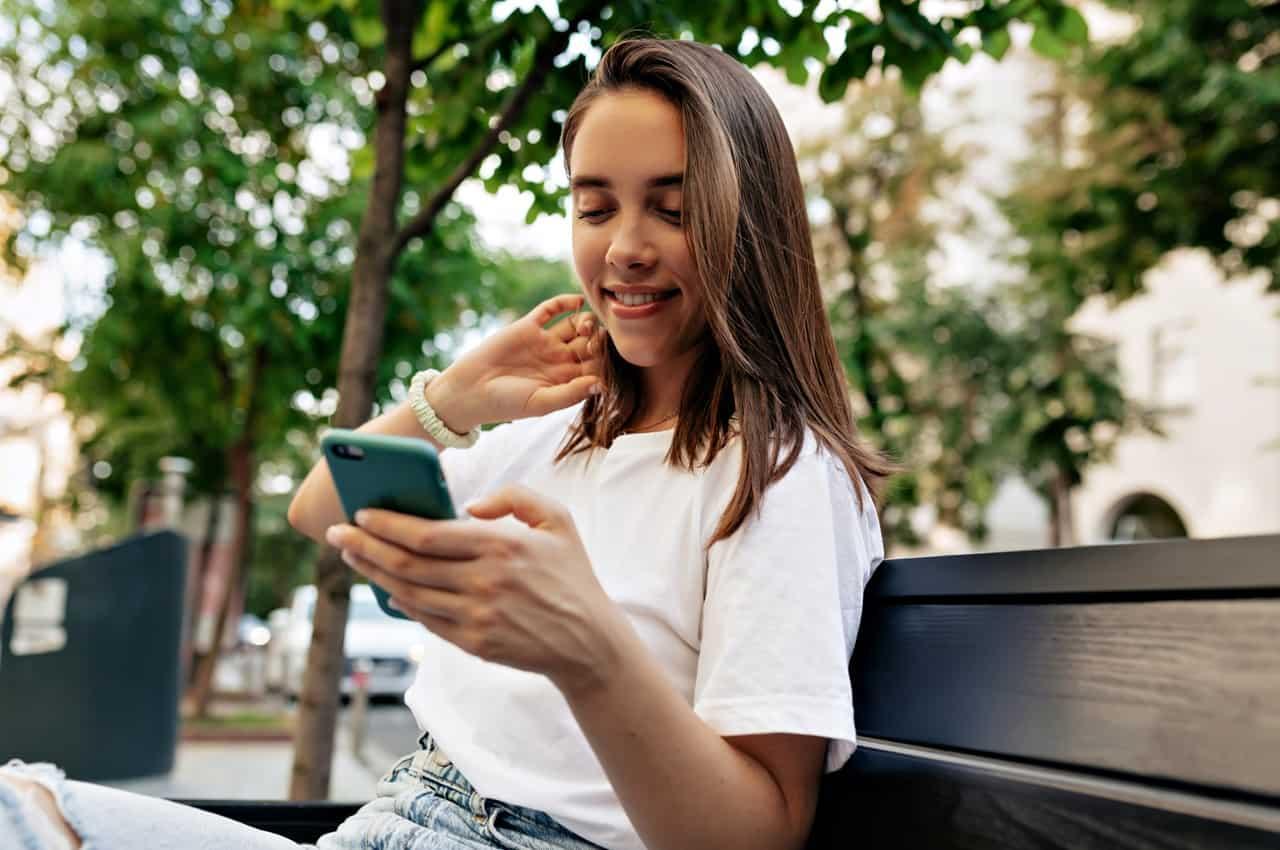 Mulher sorridente de cabelos lisos castanhos curtos, sentada no banco de uma praça e acessando empréstimo rápido e seguro no celular. A mulher veste calça jeans e blusa branca básica.