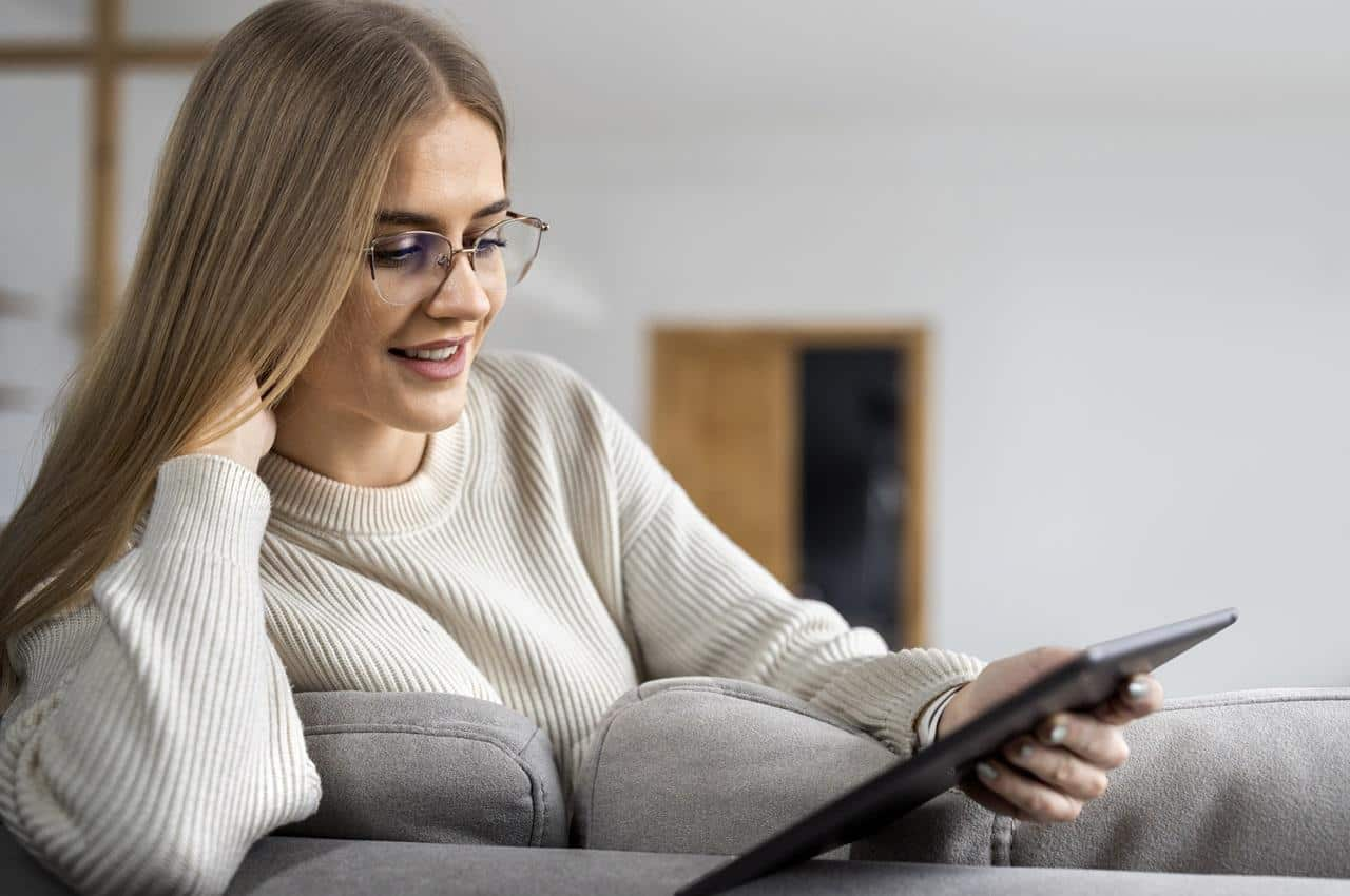 Mulher loira de óculos de grau e sorridente, vestindo um suéter creme e segurando um tablet. Ela está sentada no sofá de casa.