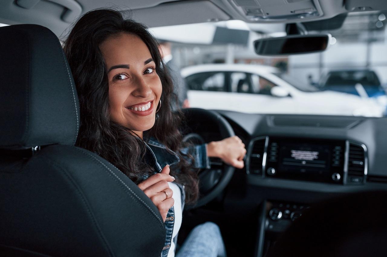 Mulher sorridente de cabelos castanhos longos. Ela está sentada no banco de motorista de um carro e olha para atrás.