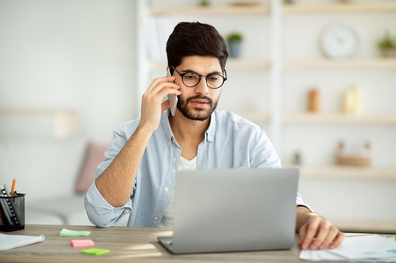 Homem com expressão concentrada, sentado em mesa de escritório e falando ao celular, enquanto acessa laptop.