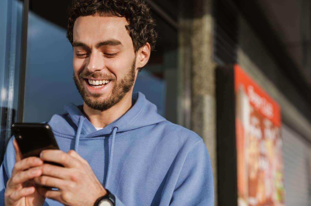 Homem sorridente usando moletom azul e digitando no celular. Ele está na calçada.