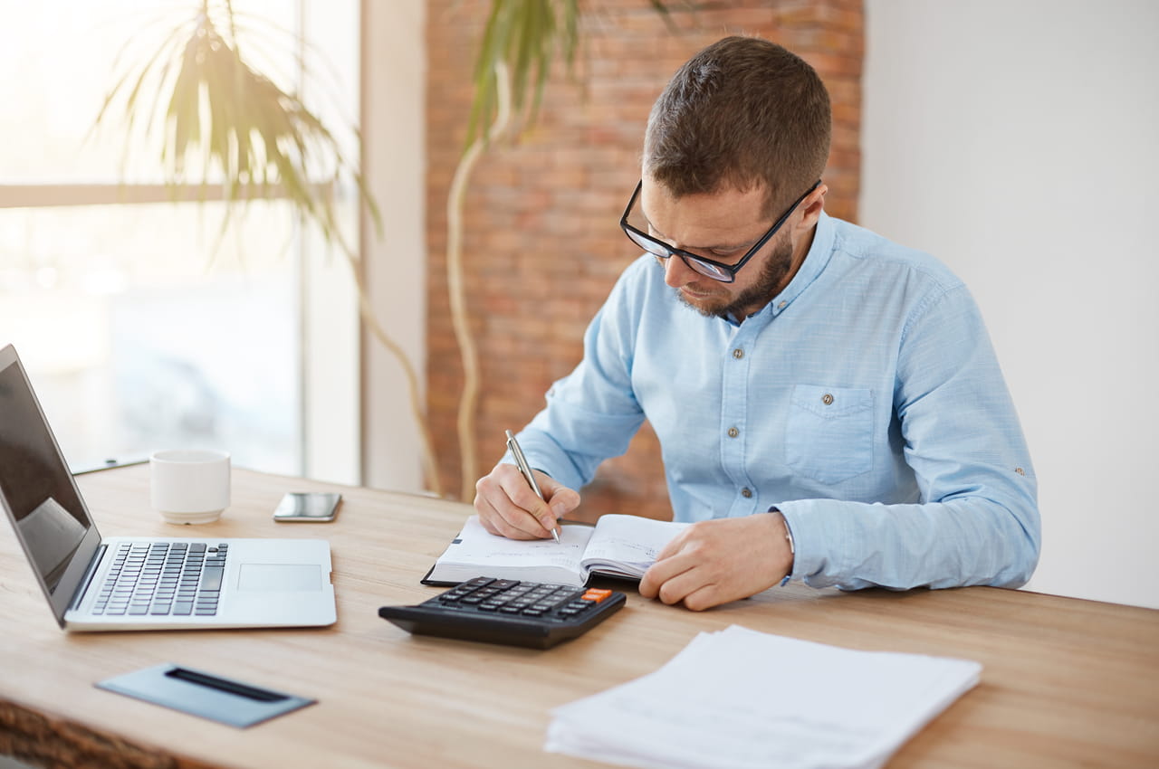 Homem com expressão concentrada, óculos de grau preto e camiseta social azul, calculando taxa pré e pós fixada de empréstimo na calculadora e agenda.