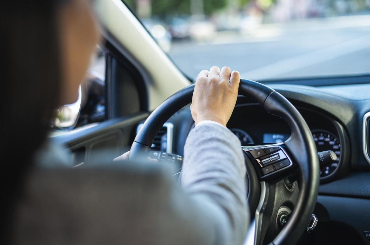 Pessoa (somente suas costas aparecem na imagem) dirigindo um carro. A imagem foca no volante do veículo.